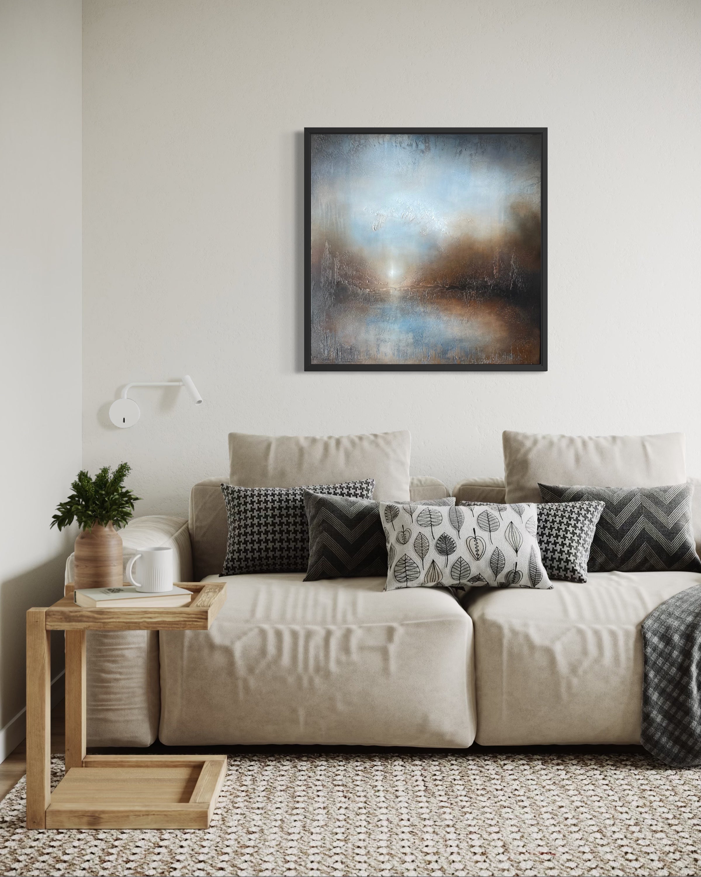A framed abstract landscape painting in muted blue, gray, brown, and cream tones hangs on a light beige wall above a cream sofa with patterned pillows in a styled living room.