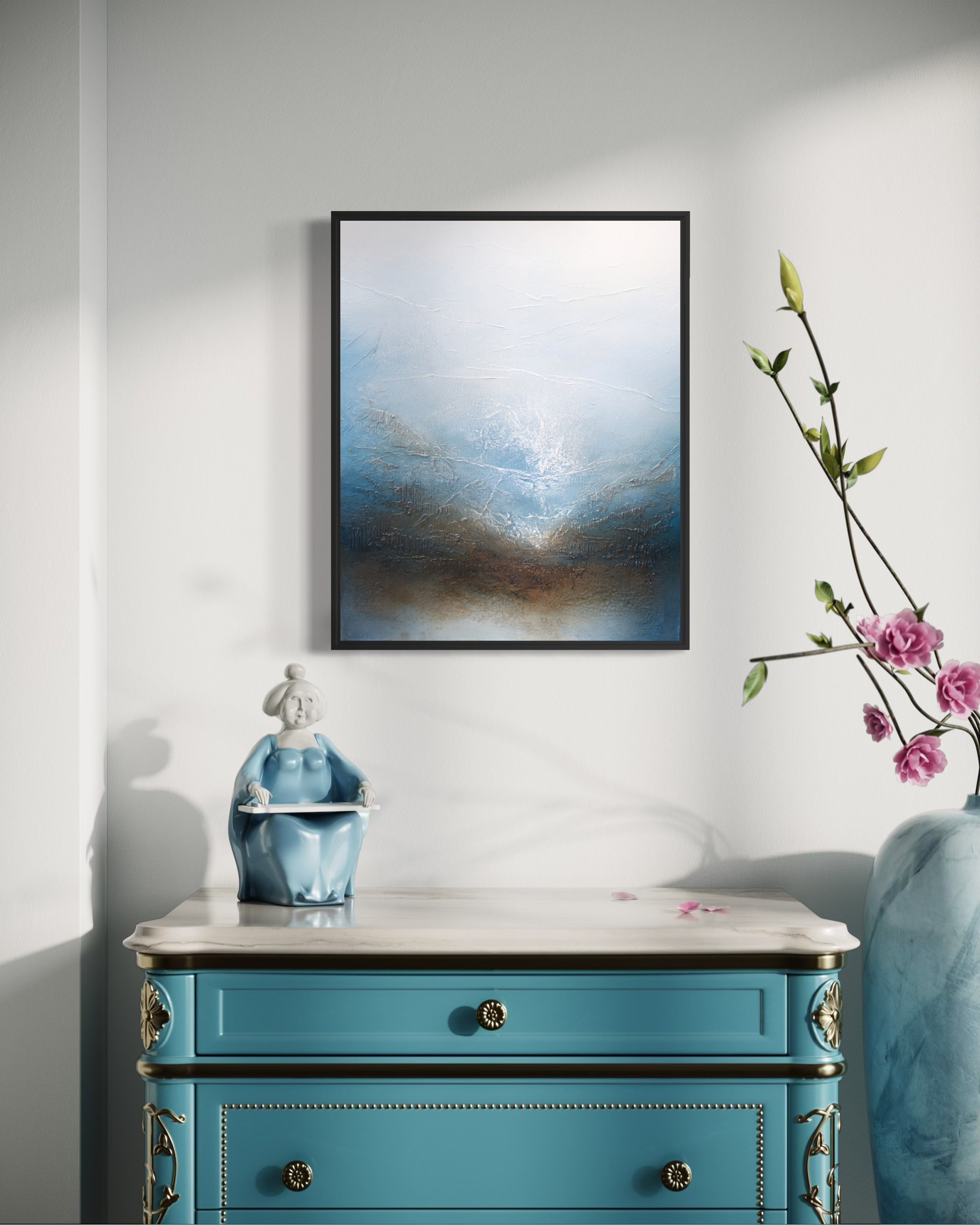 A framed abstract canvas print with blue, white, and brown tones hangs on a light gray wall above a turquoise cabinet, styled with a small figurine and a vase of pink flowers.