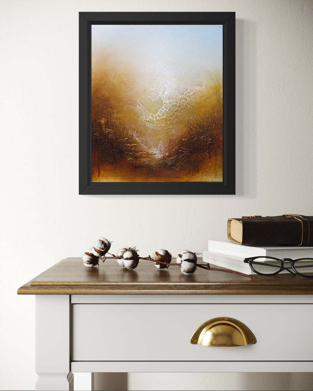 A framed abstract painting with a black frame hangs on a white wall above a wooden desk with books and cotton bolls.
