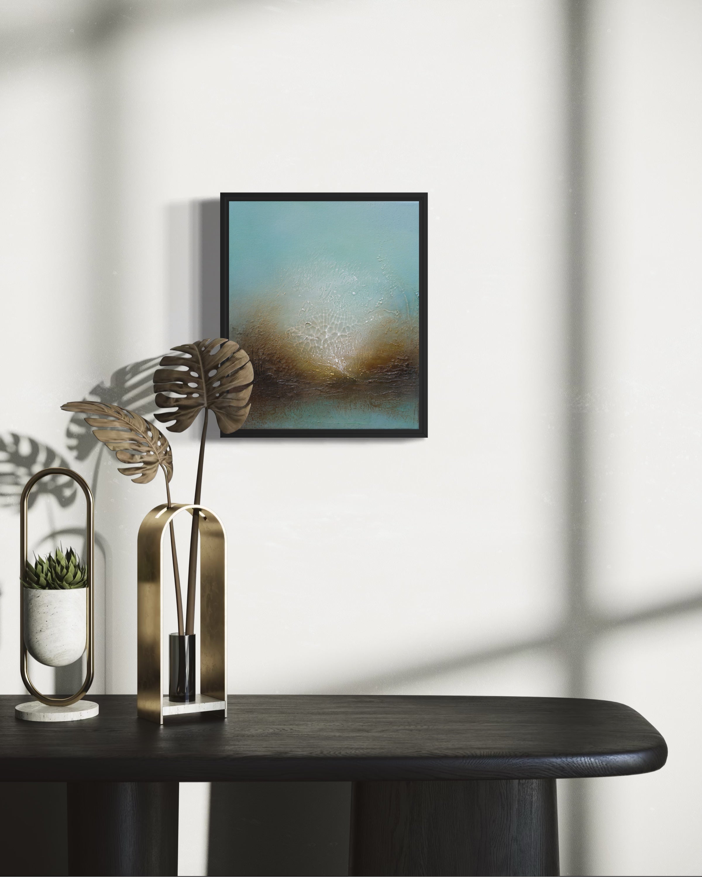 A framed abstract landscape art print with a black frame hangs on a white wall above a dark wooden console table. The artwork features a textured blue and brown scene.