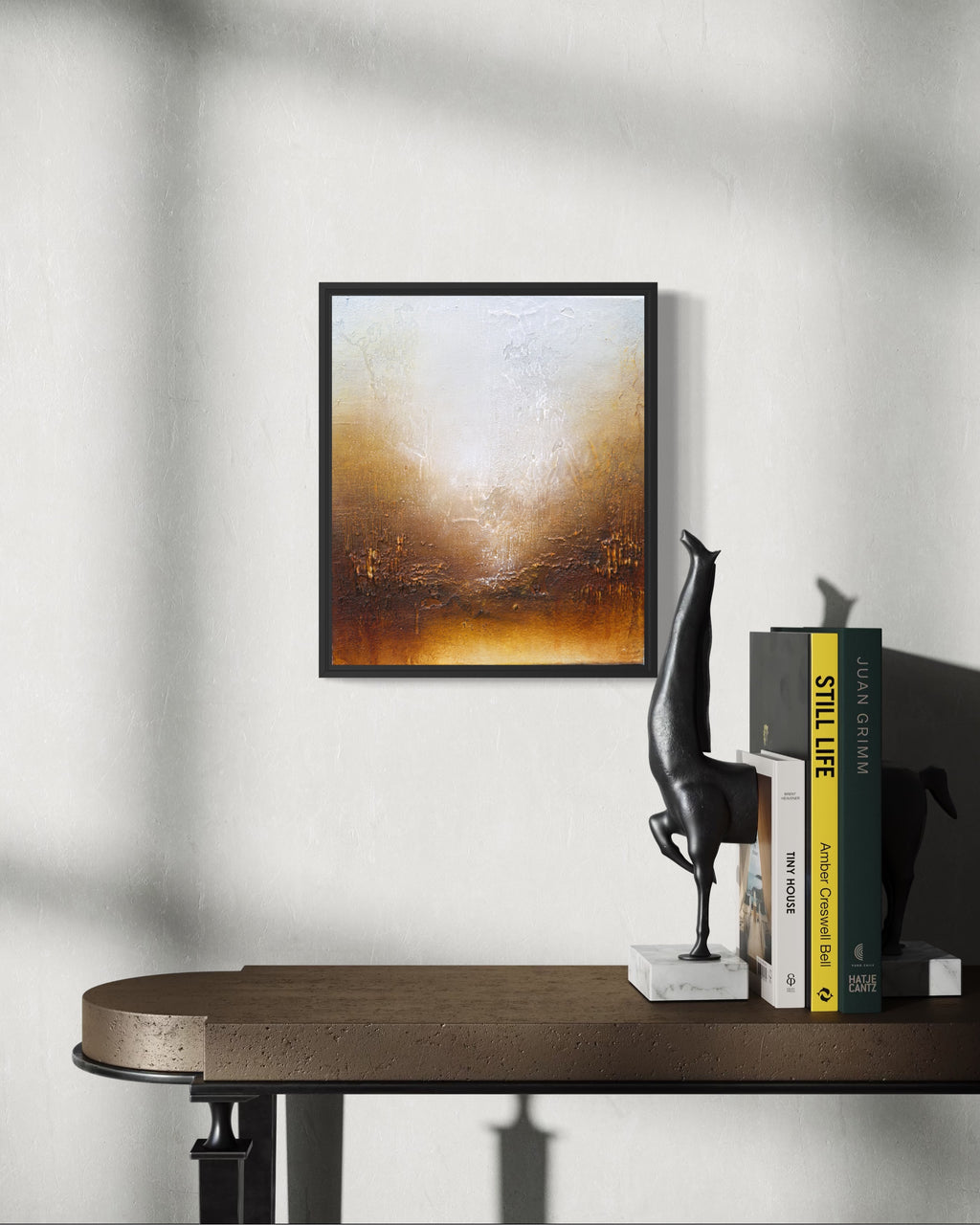 A framed abstract painting with a black frame hangs on a light gray wall above a wooden console table. The painting features a textured gradient from white and beige at the top to warm brown and orange at the bottom.