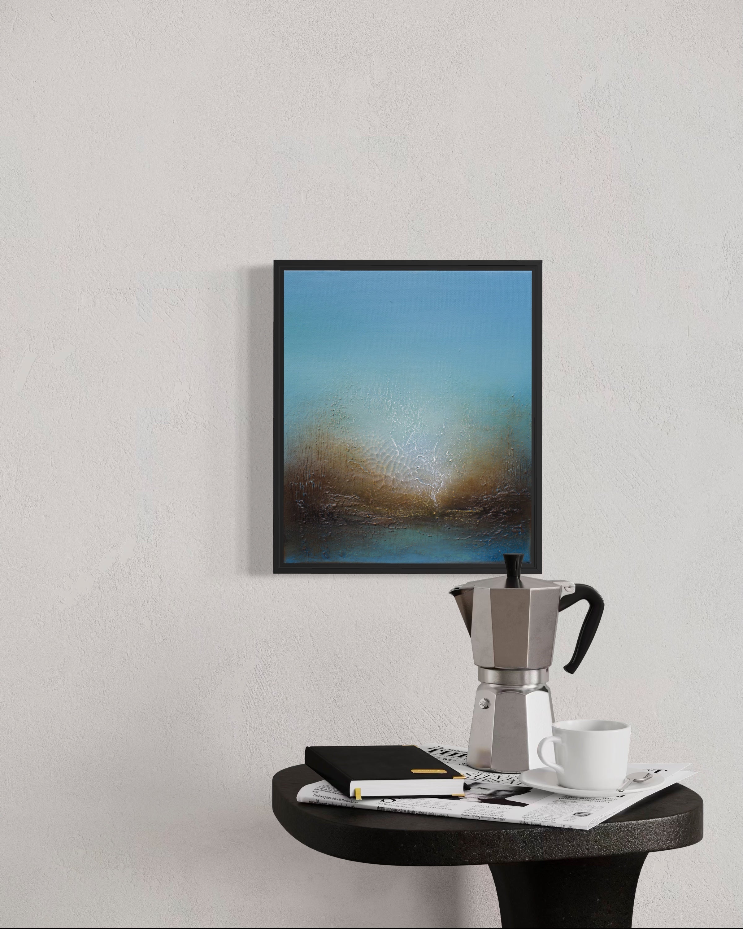 A framed abstract landscape art print featuring blue and brown tones, displayed on a textured light gray wall above a dark round table with a silver moka pot, white cup, and papers.
